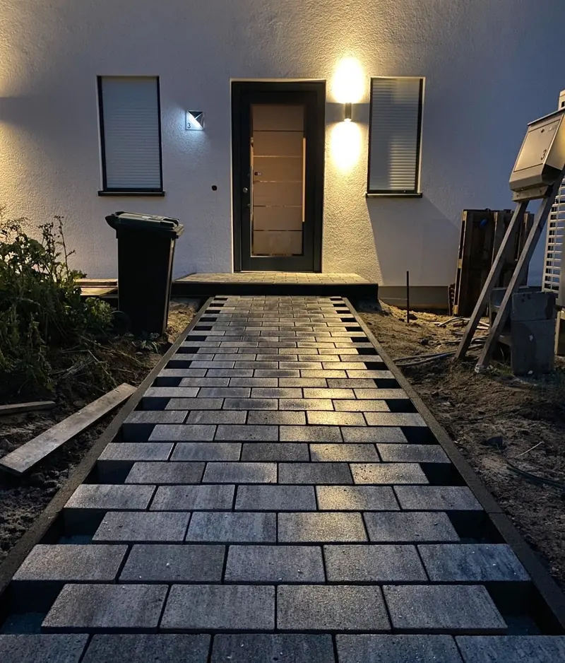 Neu gepflasterter Hauszugang aus großformatigem Betonpflaster mit dunkler Randeinfassung, abends beleuchtet
